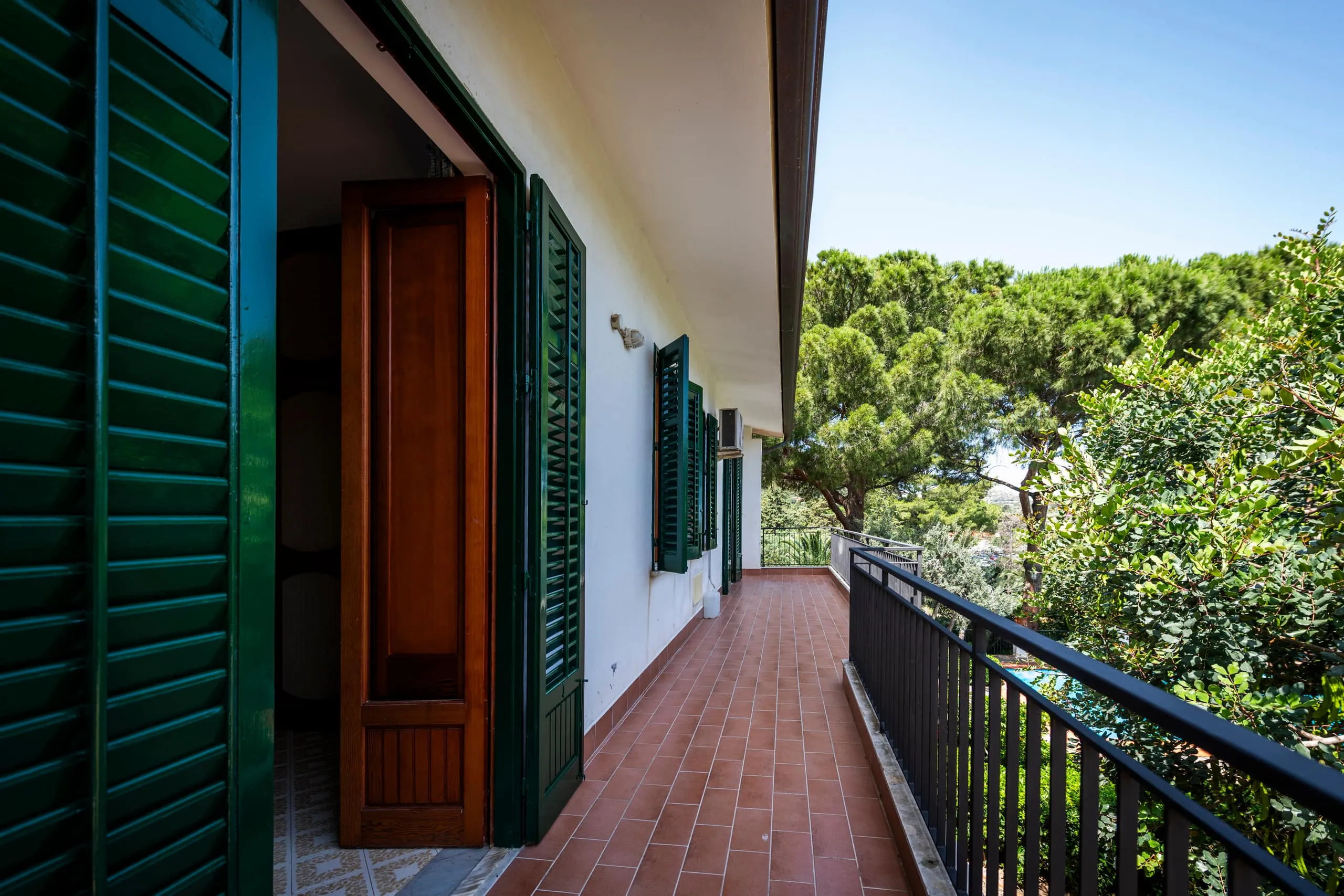 Balcone lungo in cotto con persiane verdi affacciato sugli alberi